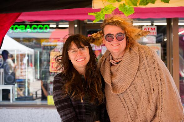 On the left a woman with a light wooly jacket and long brown hair and on the right a woman witha alight brown sweater, reddish-blonde hair with long horizontal animal ears, behind that storefronts with signs for TOBACCO and ATM