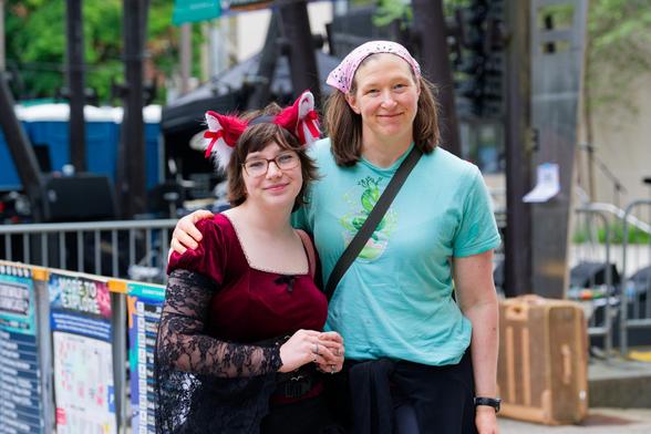 Younger woman on left with red and white animal ears and red top with lace sleeves and older woman on white with bandanna and blue-green t-shirt with barricades and other structures behind them.