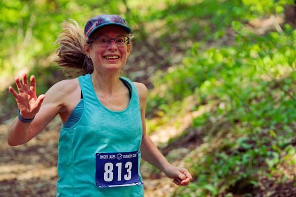 Smiling woman with flopp billed cap and pony tail trailing behind her waves to me with the ground covered with little green herbs behind her