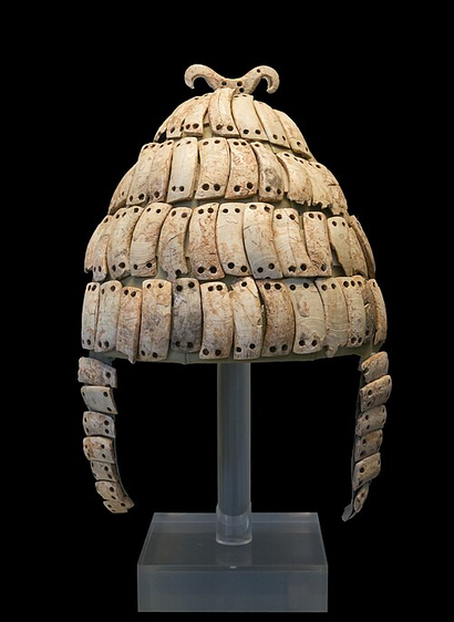 Photo of a Mycenaean Greek boar tusk helmet from Mycenae, 14th century BCE, against a black background. The individual tusk plate can easily be made out. It has a cap-like part and cheek guards. The conical shape helps deflect projectiles. It's on display at the National Archaeological Museum in Athens.