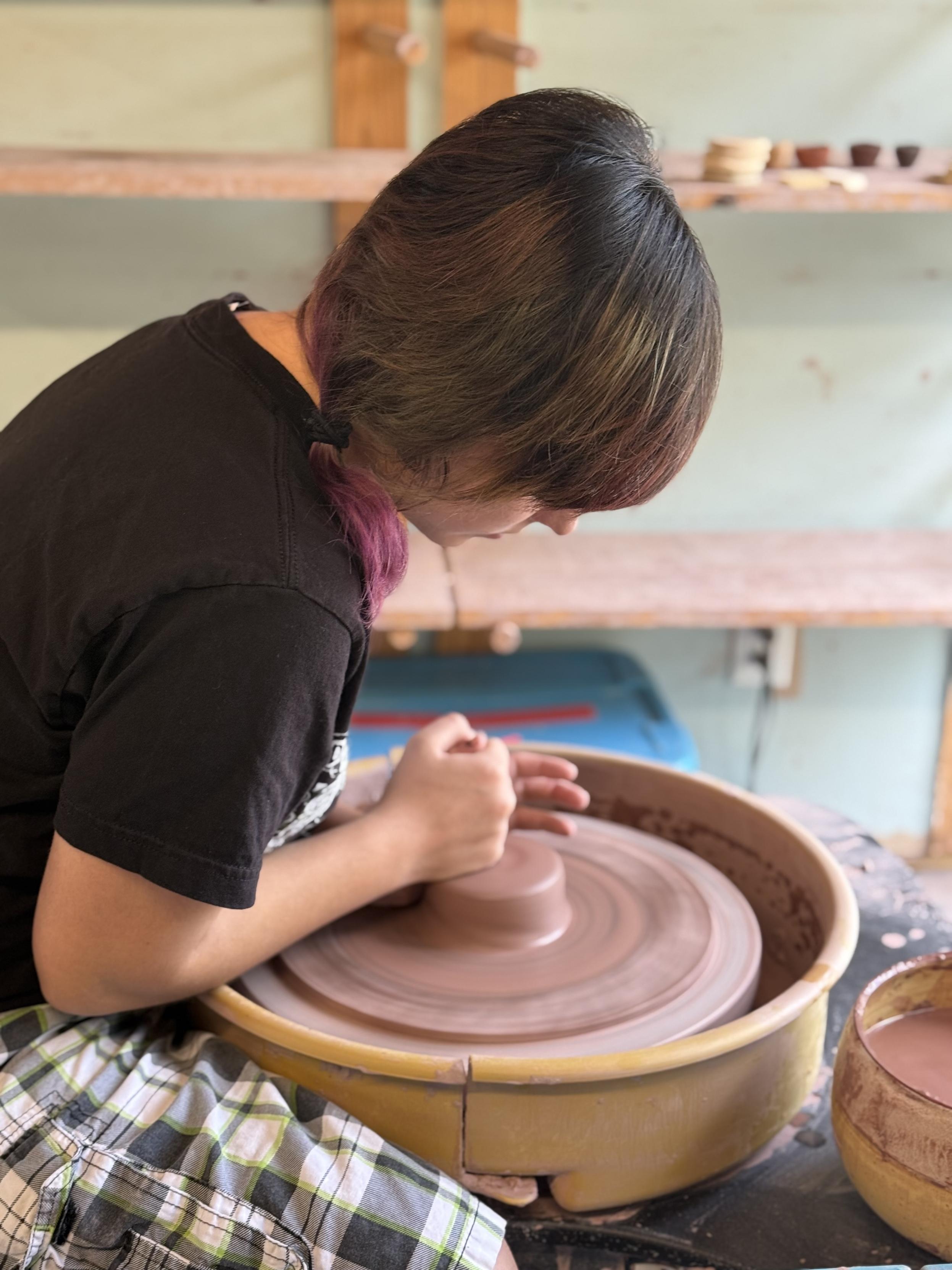 A young person with brown and purple hair wearing a black t-shirt bent over a pottery wheel, forming a lump of clay. A light green wall and wooden shelves can be seen in the background.