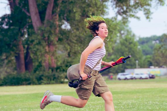 Woman running holding bag and rainbow umbrella makes eye contact with her hair flying up behind her with a few cars and a massive cottonwood tree covered with vines behind her.