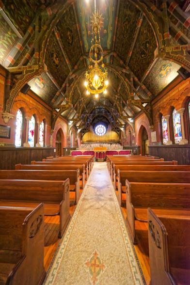 A path runs up the center past multiple rows of short pews and terminates in an altar with numerous votive candles and a circular stained glass windows:  the walls on both sides have arch-topped stained glass windows and the ceiling has elaborate paintings and a chandelier.