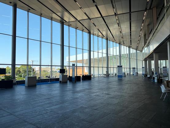 A view of a large, empty conference center hall. . There are floor-to-ceiling windows along the back wall with blue sky outside. The floor looks like a marble and the ceiling is metal with slots in it. 