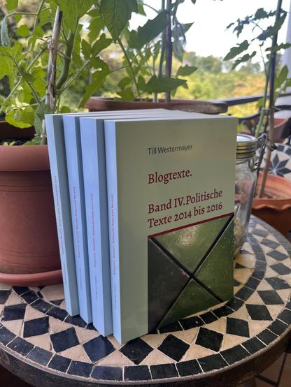Vier helltürkisfarbene Bücher (Blogtexte I-IV) stehen vor einem Blumentopf auf einem Mosaiktisch. Auf dem vorderen Band (IV) ist ein Foto mit verschiedenen Grüntönen zu sehen.