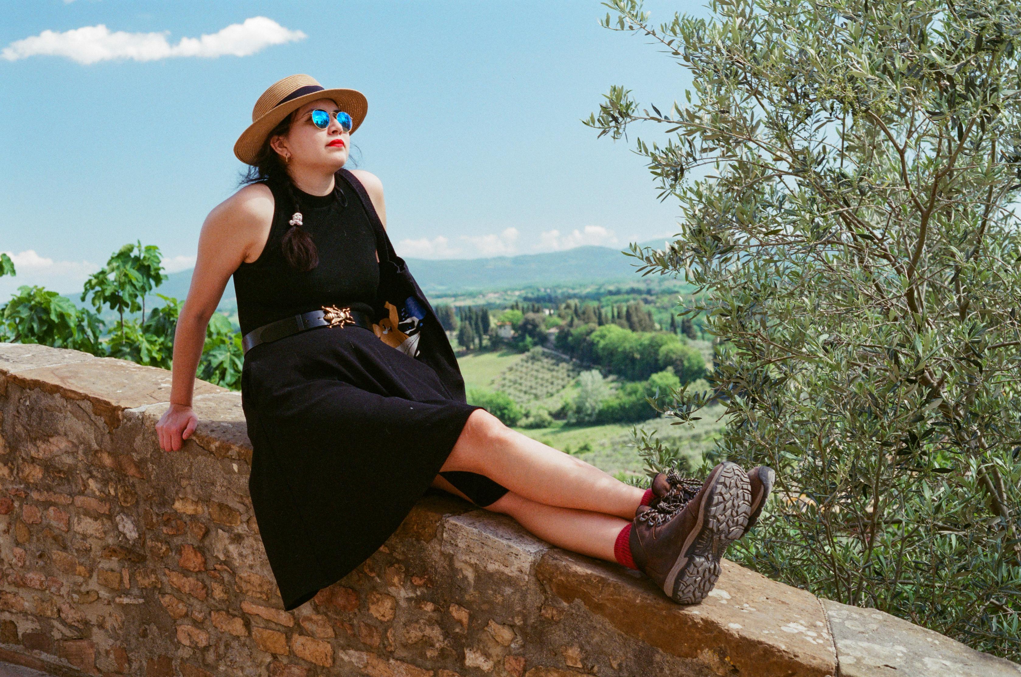 A beautiful woman in a black sleeveless top and a knee-long black skirt, with a straw hat and a belt featuring a brass bee, wearing red lipstick and reflective sunglasses, sitting on the ledge of a beige stone wall overseeing a green landscape filled with cypress trees under a pastel blue sky.