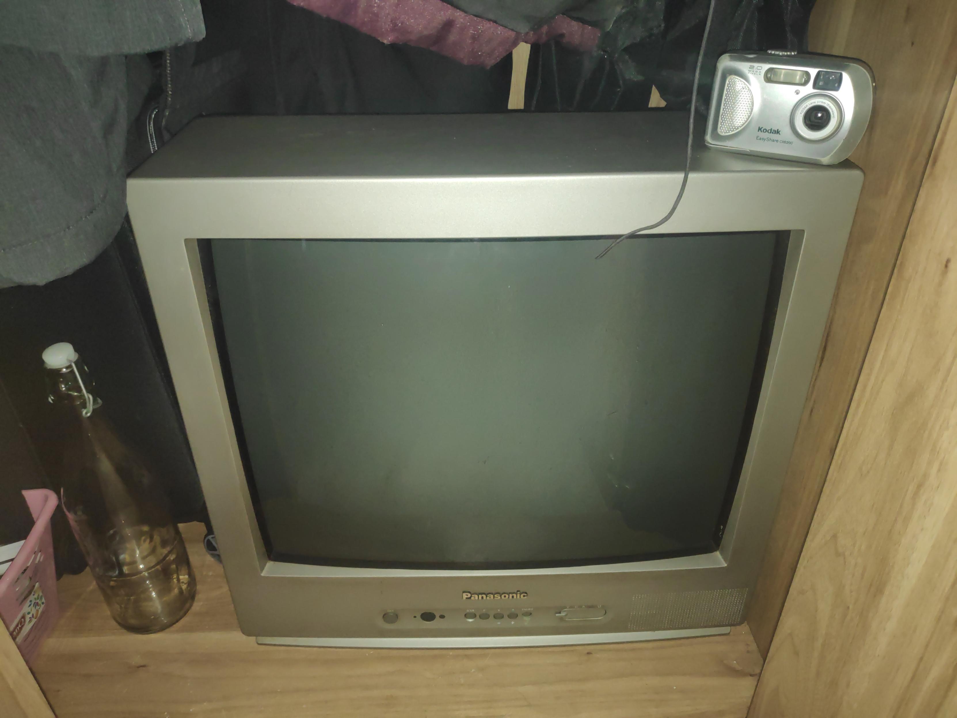 A photo of a grey Panasonic TV inside a wardrobe full of clothing. It has a Kodak camera on top of it and an empty glass bottle next to it.