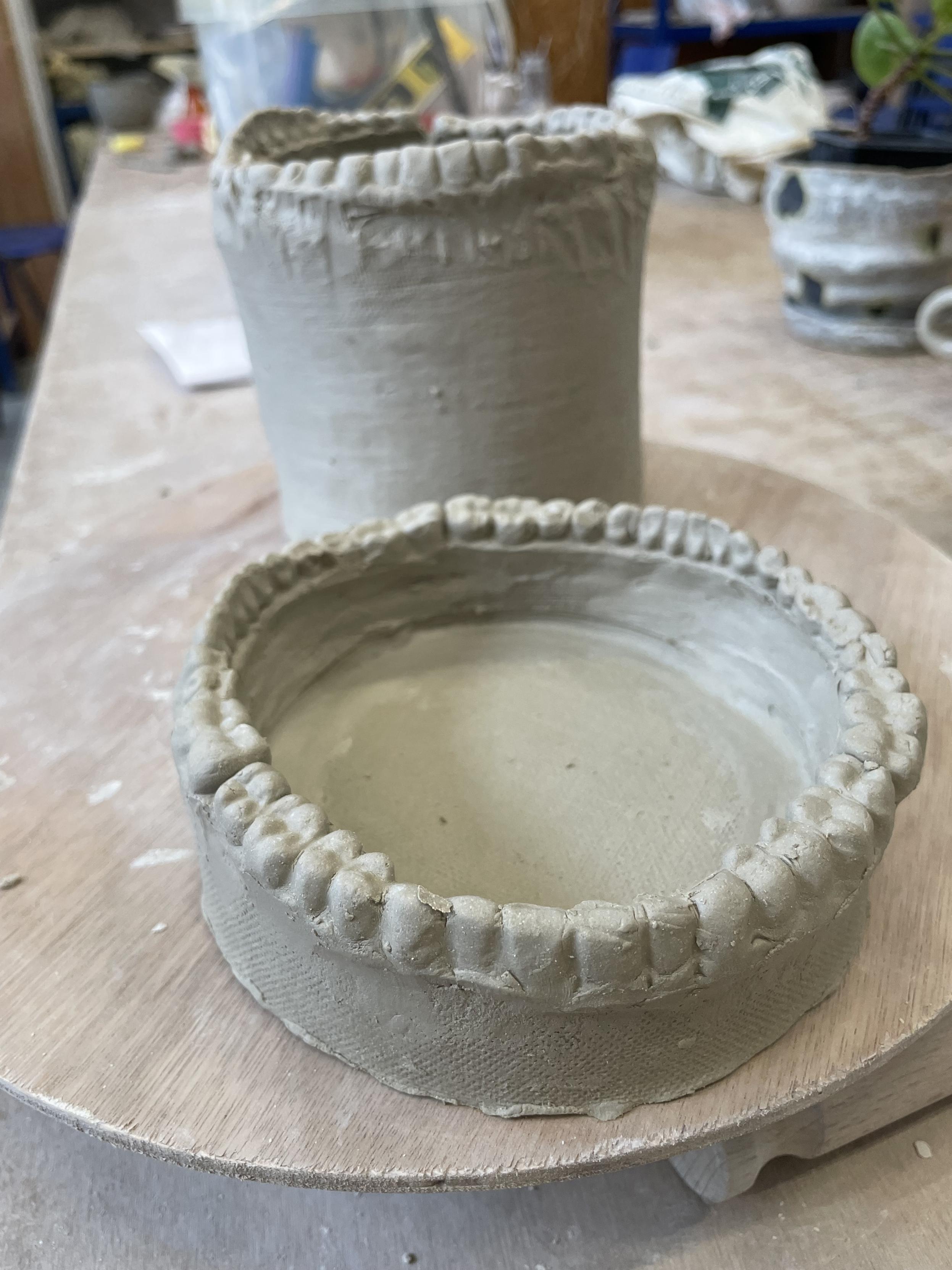 Two work in progress pottery vessels, a taller mug and shorter dish, both rimmed with moulded human teeth. 