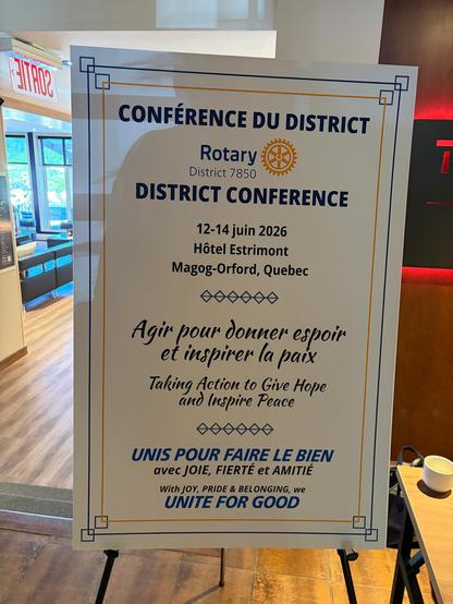 A large white sign in a hotel lobby that says in English and French “District Conference” , the date, location, and Rotary slogans