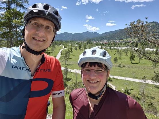 The image features two cyclists taking a selfie during their ride. Both are wearing helmets for safety. The person on the left is dressed in a vibrant cycling jersey with red, white, and blue colors, and the word "PROTECTIVE" is visible on it. The person on the right is wearing a maroon jersey. Both individuals are smiling, radiating joy and enthusiasm. Their attire suggests they are prepared for a serious cycling session, emphasizing both functionality and style. The helmets and jerseys indica…