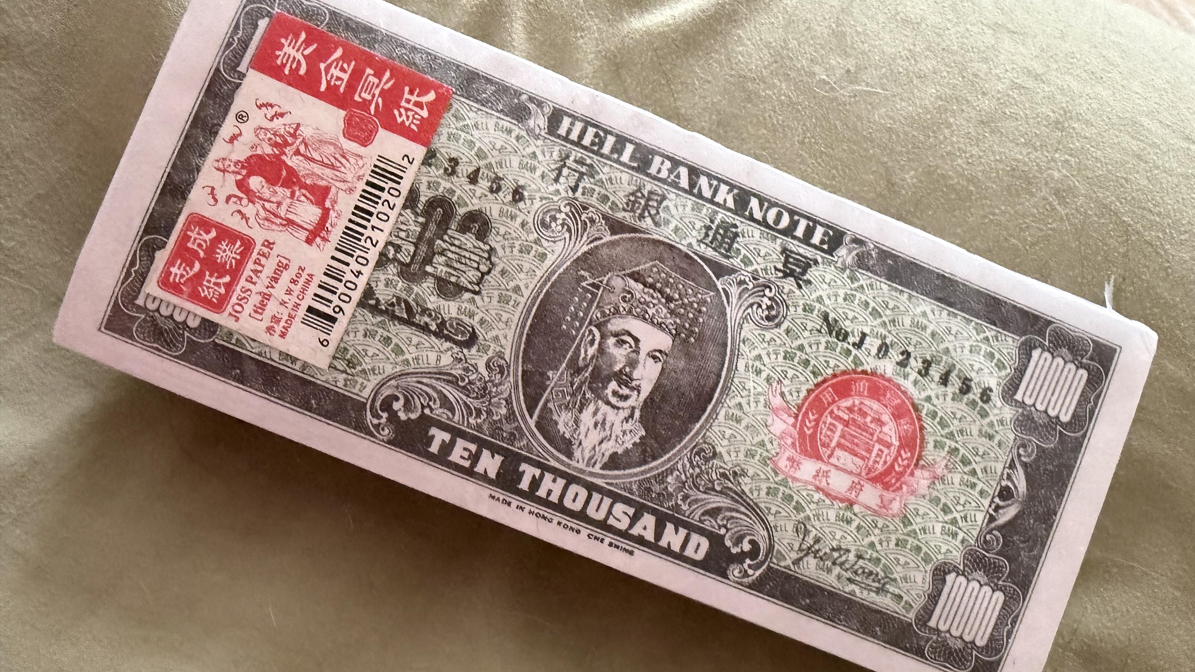 A decorative Hell Bank note with a denomination of ten thousand. The note features an image of a figure wearing traditional Chinese attire, along with Chinese characters and a red stamp. It is placed on a soft, light-colored surface.