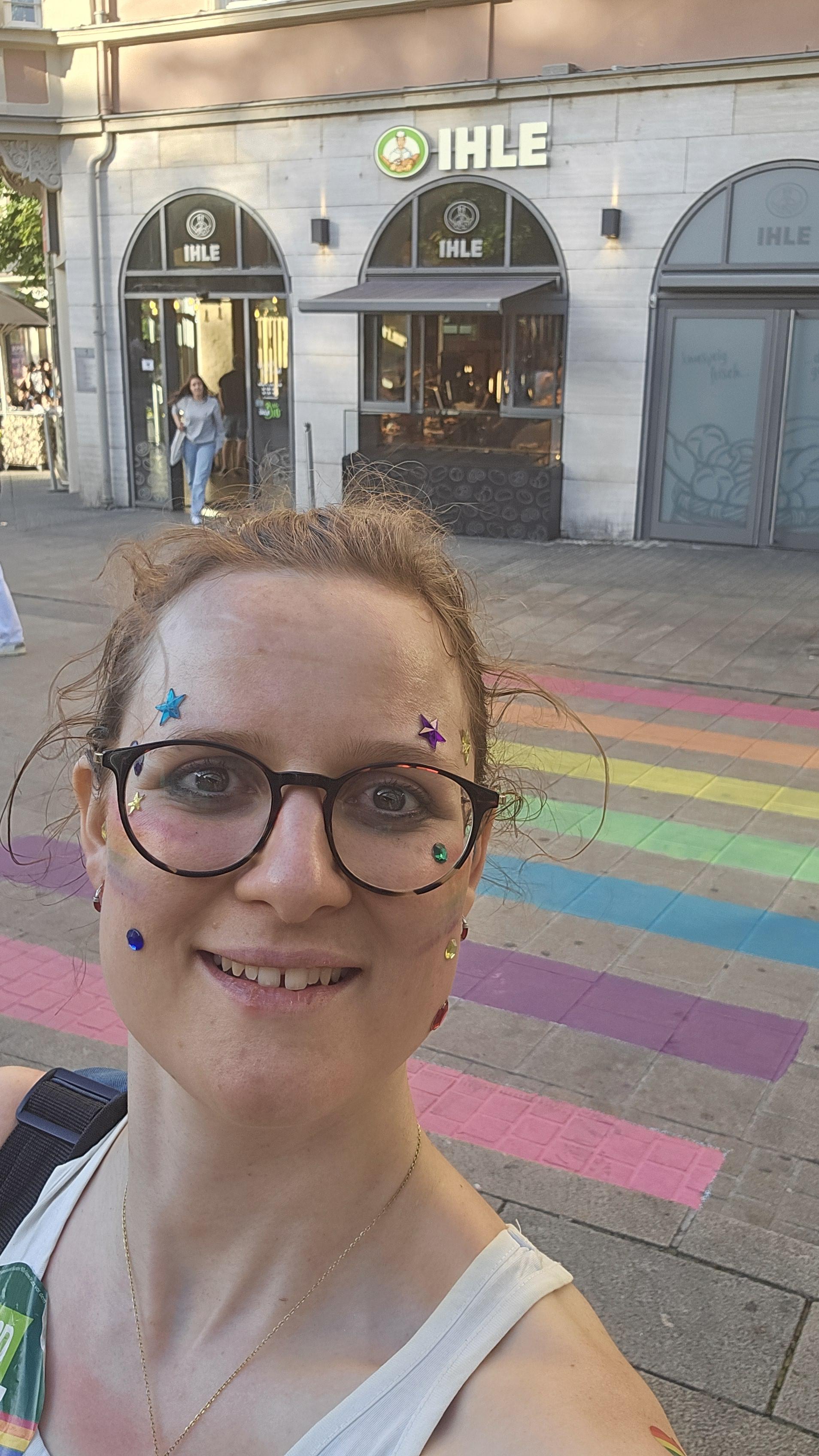 Ein Foto von mir mit Glitzersteinen undRregenbogenfahne im Gesicht vor einem Regenbogenzebrastreifen am Augsburger Königsplatz.