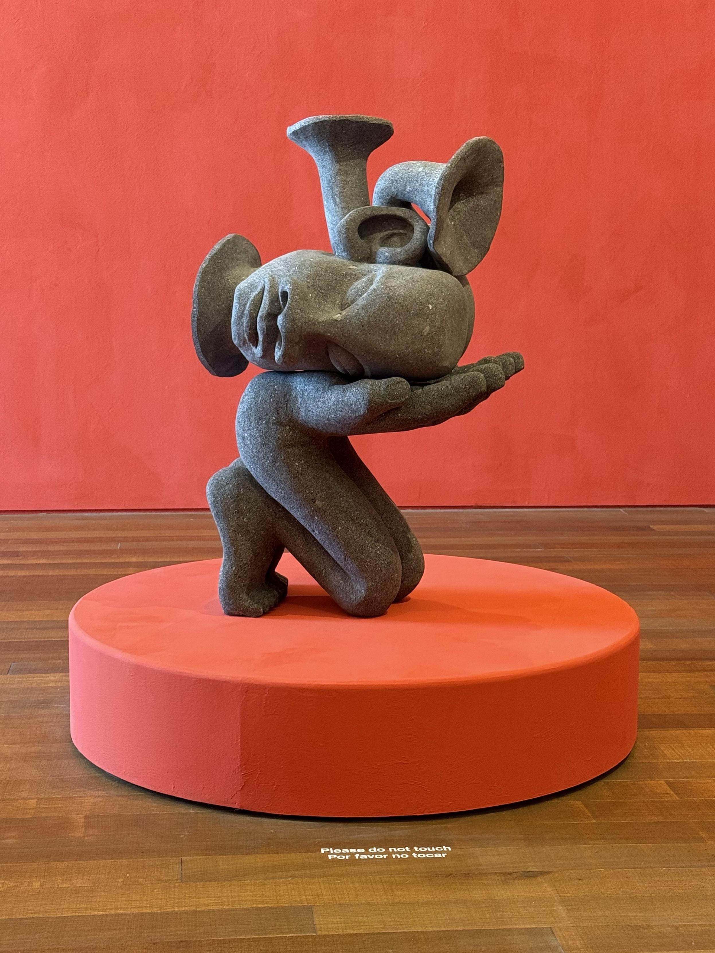 A stone sculpture of a stylized figure kneeling, holding a head with exaggerated features. The sculpture is mounted on a round red pedestal, against a matching red wall in the background.
