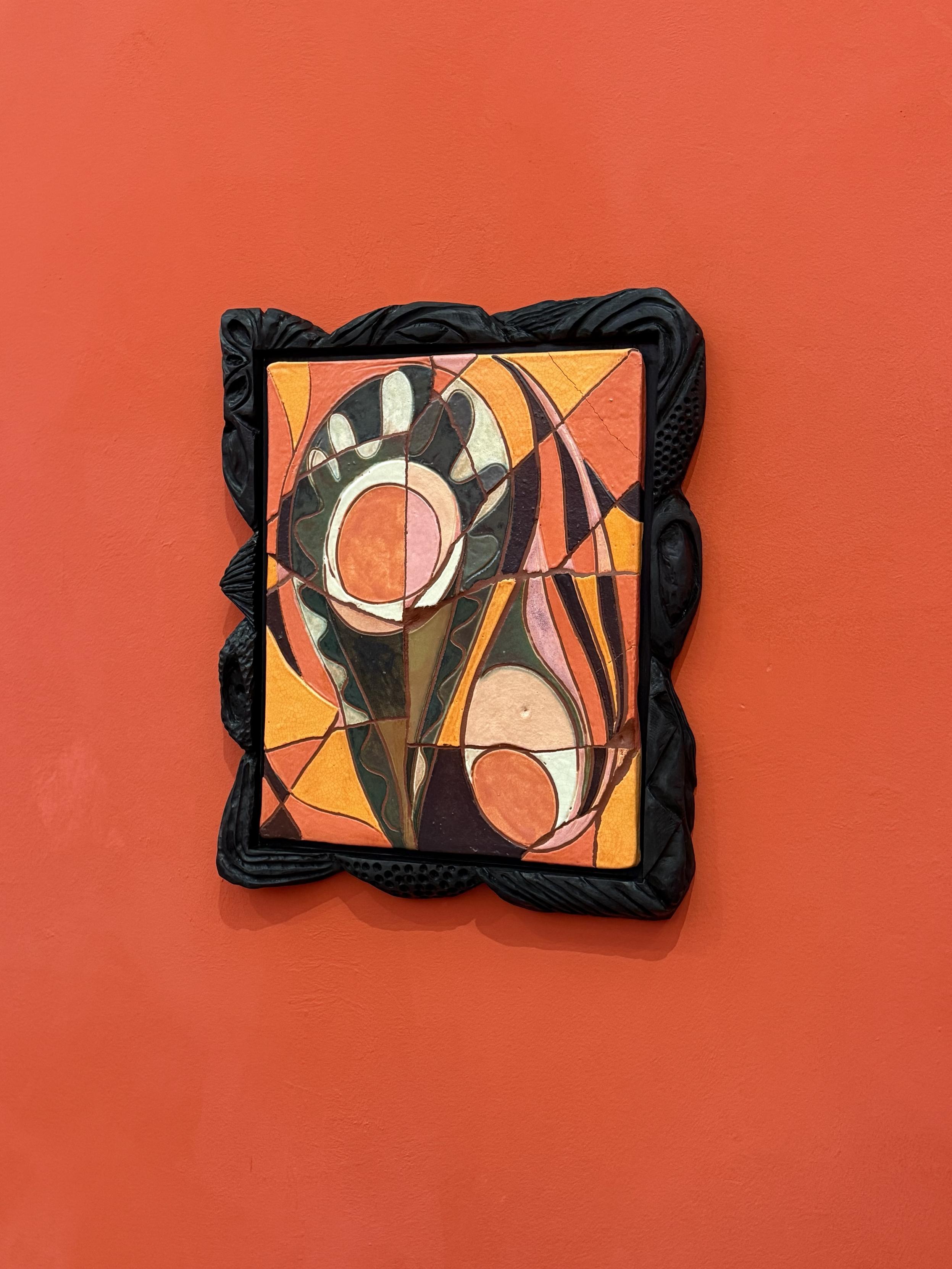 An abstract ceramic mosaic artwork framed in ornate black wood, featuring geometric shapes and vibrant colors like orange, green, and brown. The background is a solid red-orange wall.