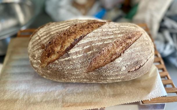 Ein tief diagonal eingeschnittenes, längliches Brot auf einem Holzgitter und Backtrennpapier am Auskühlen. Das Brot hat helle Längsstreifen aus hellerem Mehl von der Gärung im Bast-Gärkörbchen.