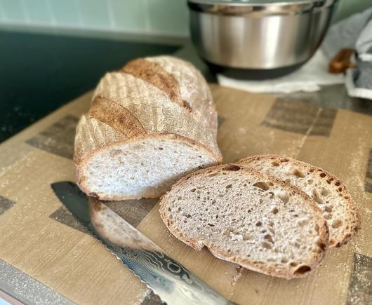 Längliches Brot mit Schnittfläche zu mir. Daneben liegen zwei grob geporte Schnitten. Alles liegt auf Brotpapier. Vorne sieht man das schön gemusterte Brotmesser aus Damast.