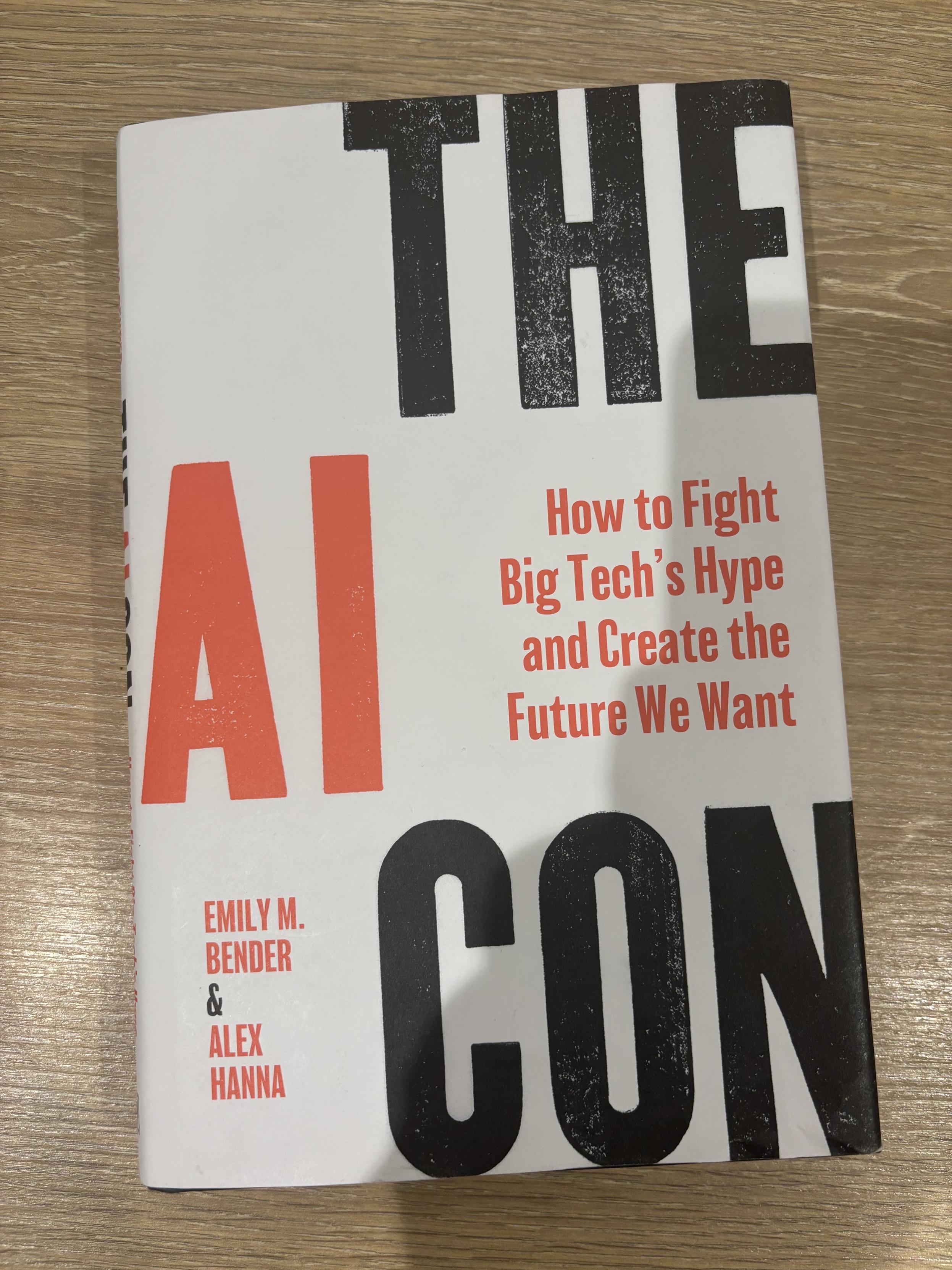 A book laying on a wooden table, white cover with large words in black and red: The AI Con - How to fight big tech’s hype and create the future we want.
