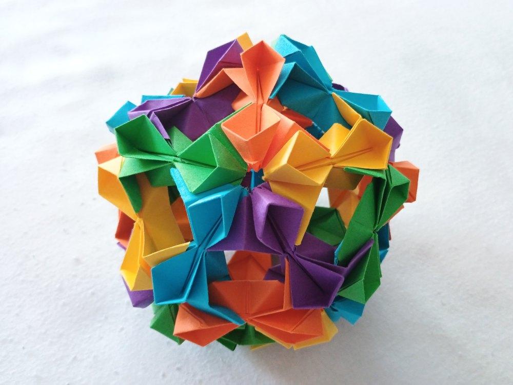 Modular origami icosahedron made out of flowery edge pieces in blue, green, yellow, purple, and orange. Photo against a white background.
