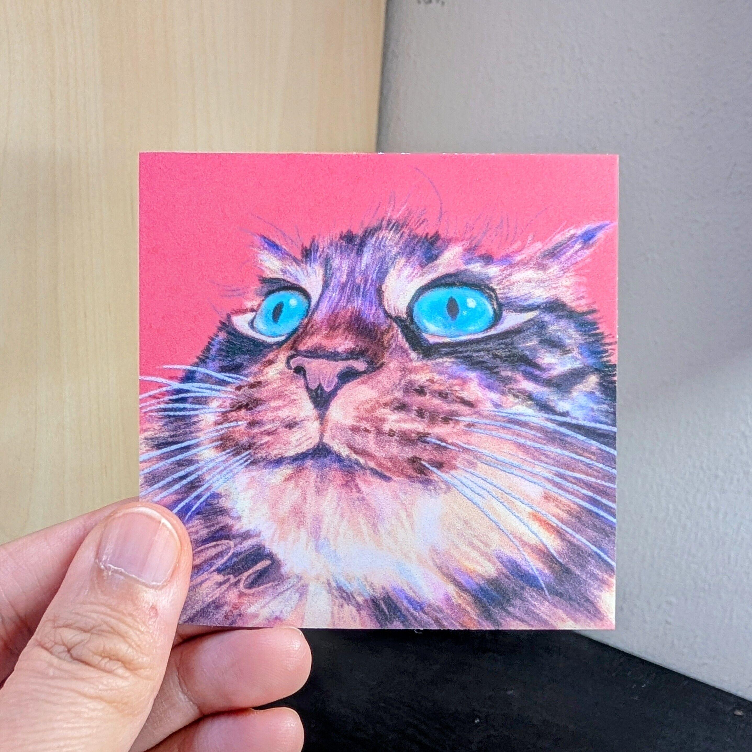 Photo of a left hand holding a square sticker. The sticker is a matte painting of a cat with teal eyes.