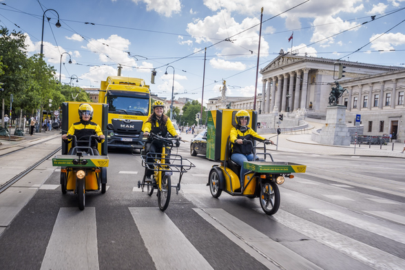 Yellow postal service electric tricycles and bike on a Vienna street, followed by a big yellow truck