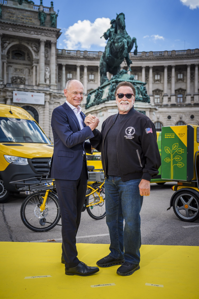 2 persons arm wrestling in front of yellow postal bikes and vans, and statue