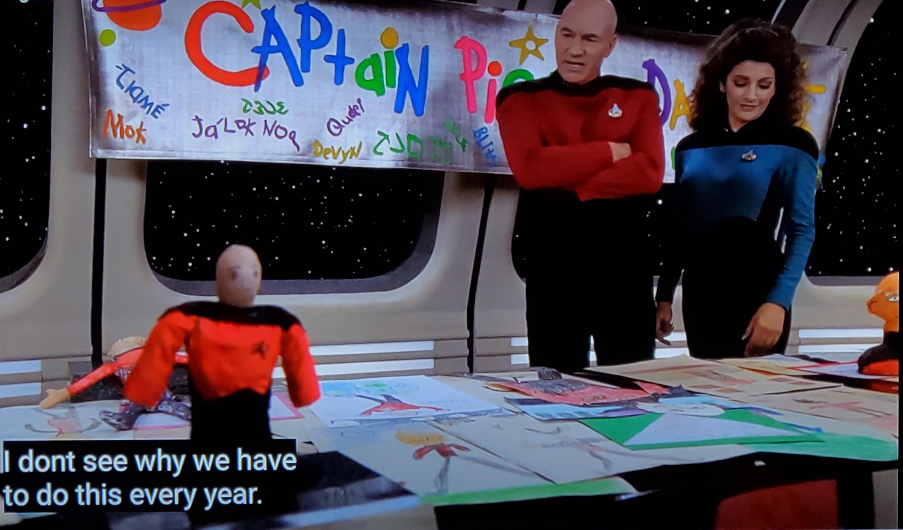 Star Trek The Next Generation scene. We're on the Enterprise, in a room with many windows out to space. It looks like a classroom type setup with a table and crafts in the background, but also probably Ten Forward with a different set. Picard is standing in front of a banner hanging in the room with 'Captain Picard Day' drawn on it. The letters are different colors and clearly drawn by children, like if you were a righty but tried to write with your left. The banner also has different cute little other drawings around the letters, like planets and names of kids. In front of Picard is a table covered in children'a drawings and two very very rough sculpturs or poorly made dolls of the Captain in uniform. His arms are crossed and he looks a teensy weensy bit irritated. Troi is next to him also regarding the pictures, but she's smiling and looks pleased. Closed caption reads, "I don't see why we have to do this every year."
