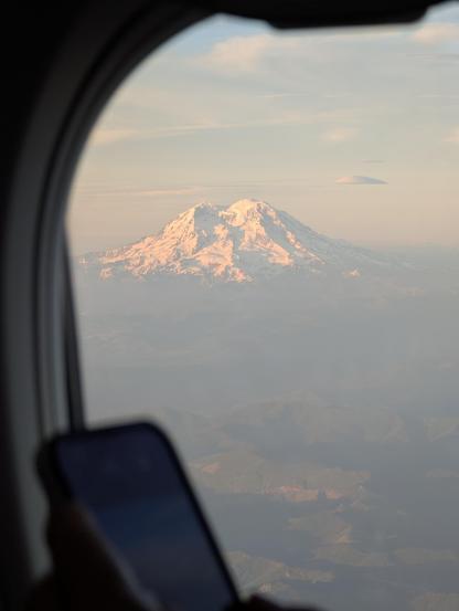 Photo of Mount Rainier through a plane window. The foreground is out of focus, showing the plane window and the phone of the person who is actually in the window seat. The snowcapped mountain sits in the center of the frame, basking in the golden orange glow of a Seattle summer sunset.