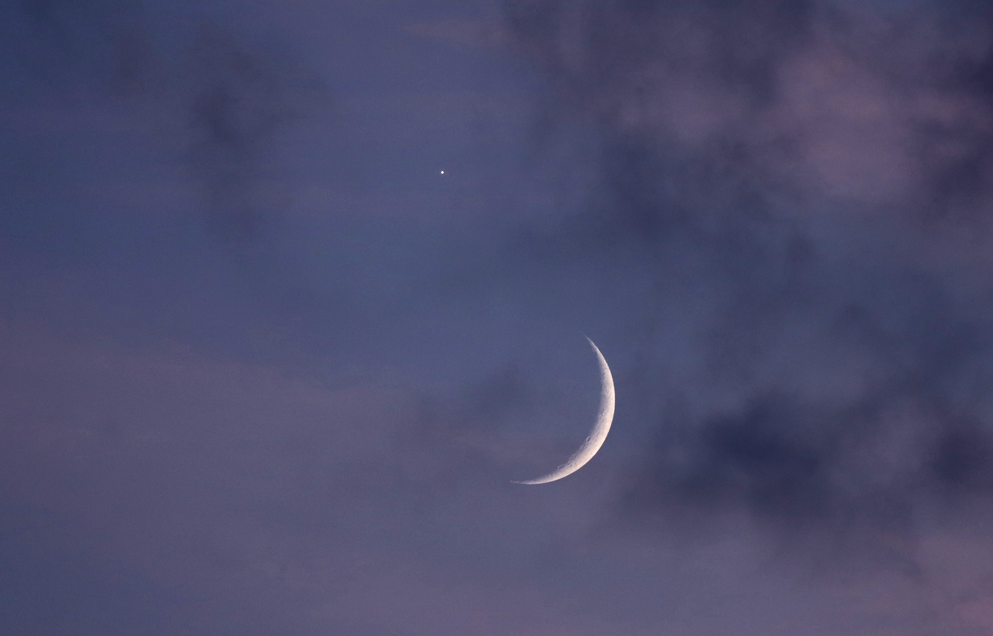 Photo of the evening sky with low and high clouds. A toenail Moon is visible in the center, with Venus very nearby, visible as a bright spot.