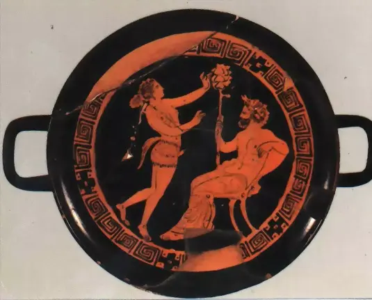 Red-figure vase painting. The god Dionysos is seated, holding his thyrsos staff. In front of him, a maenad is acting, wearing phallus trunks, a costume to depict satyrs in theatre.