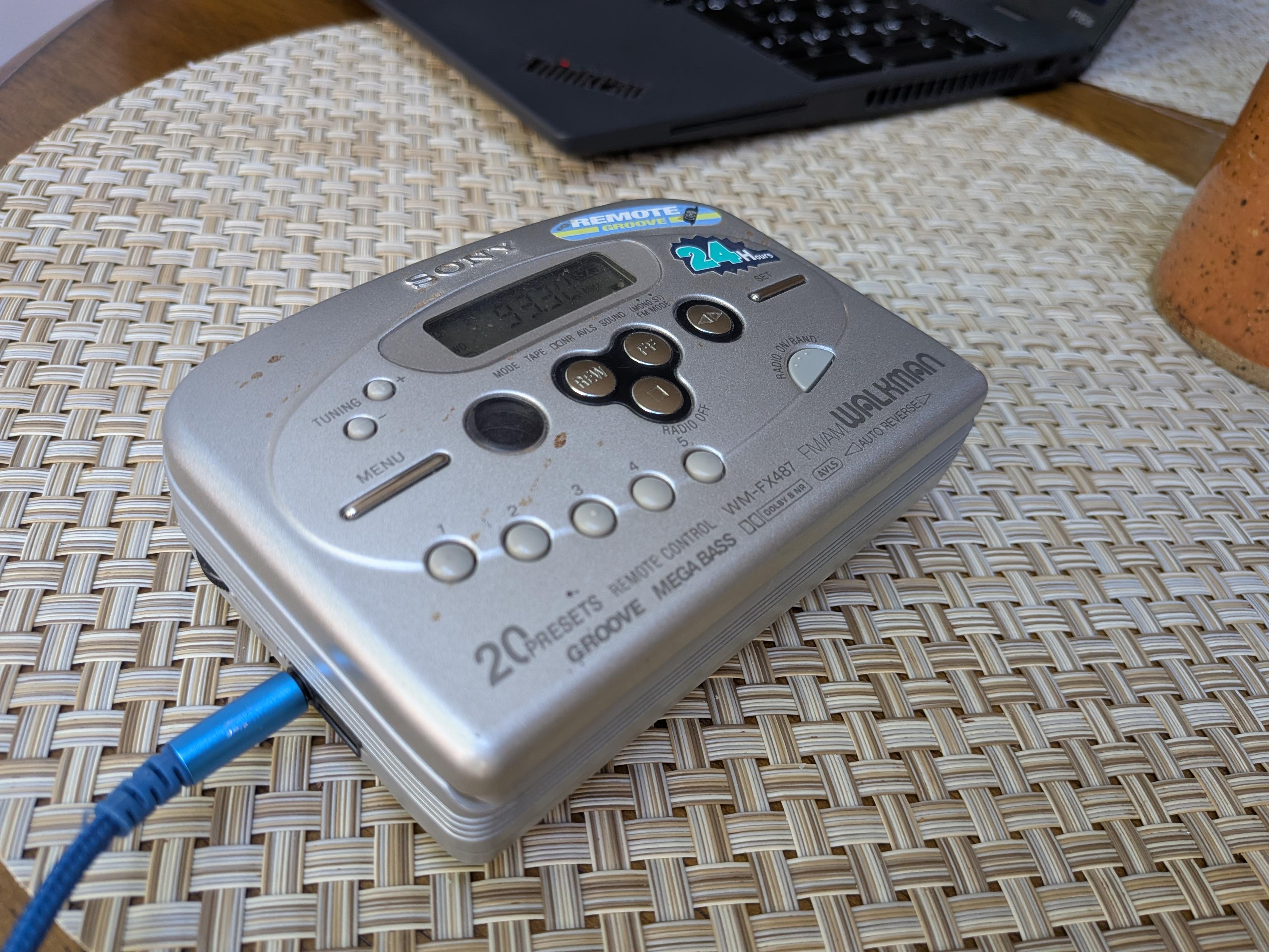 An old beat-up silver Sony Walkman. This one is from an electronic era, with an LCD display and "sensor" (but still physical) buttons. A blue 3.5mm headphone jack is inserted at the side.