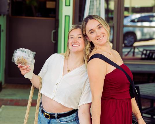 Two smiling young blonde women almost back to back,  one with a white top with an open midriff and the other with a reddish top with bare shoulders.  Now that I think of it I should have gotten one of them to put down a food container