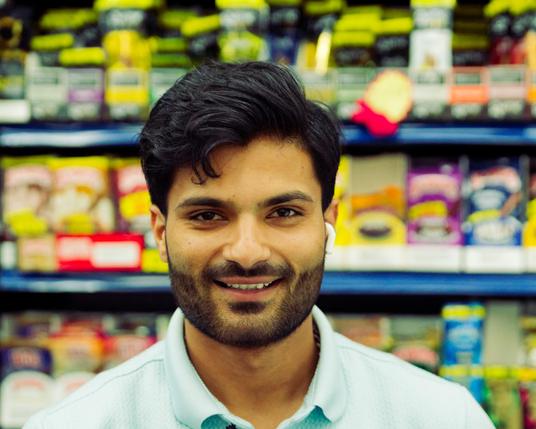Man with beard and moderate dark hair and moderately dark complexion with light blue shirt is smiling in front of blurred out tobacco products.