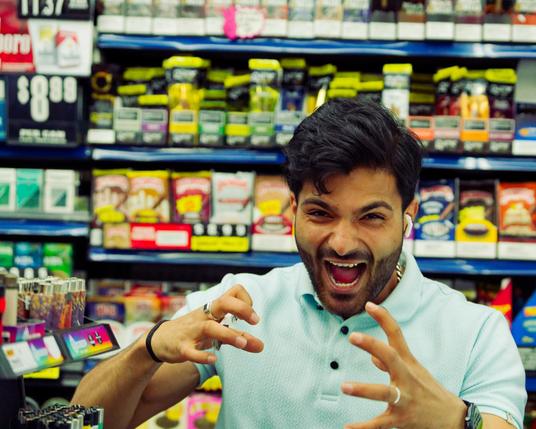 Dark haired and slightly brown man makes fierce expression in front of tobacco display