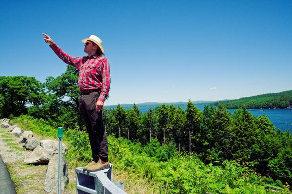 Young man with cowboy hat and plaid shirt points to upper left with lake view behind him partially obstructed by evergreen trees