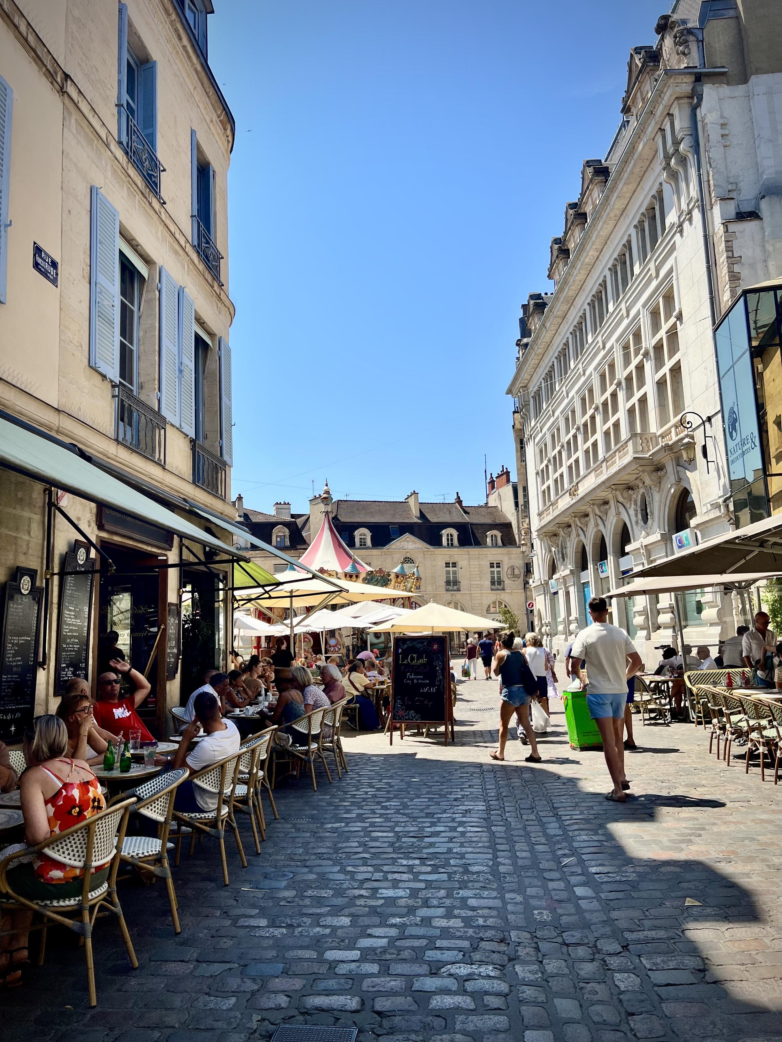 Rue Francois Rude, Dijon

A sunny outdoor street café scene in a historic town square. Tall stone buildings frame the narrow lane on both sides, with shuttered windows on the left and an arched building entrance on the right. In the foreground, café tables and chairs fill the left side where people are seated and talking. Several umbrellas—mostly white with one striped—shade more tables deeper in the scene. A cobblestone walkway runs through the middle toward the back, where more people walk and mingle under bright blue sky.