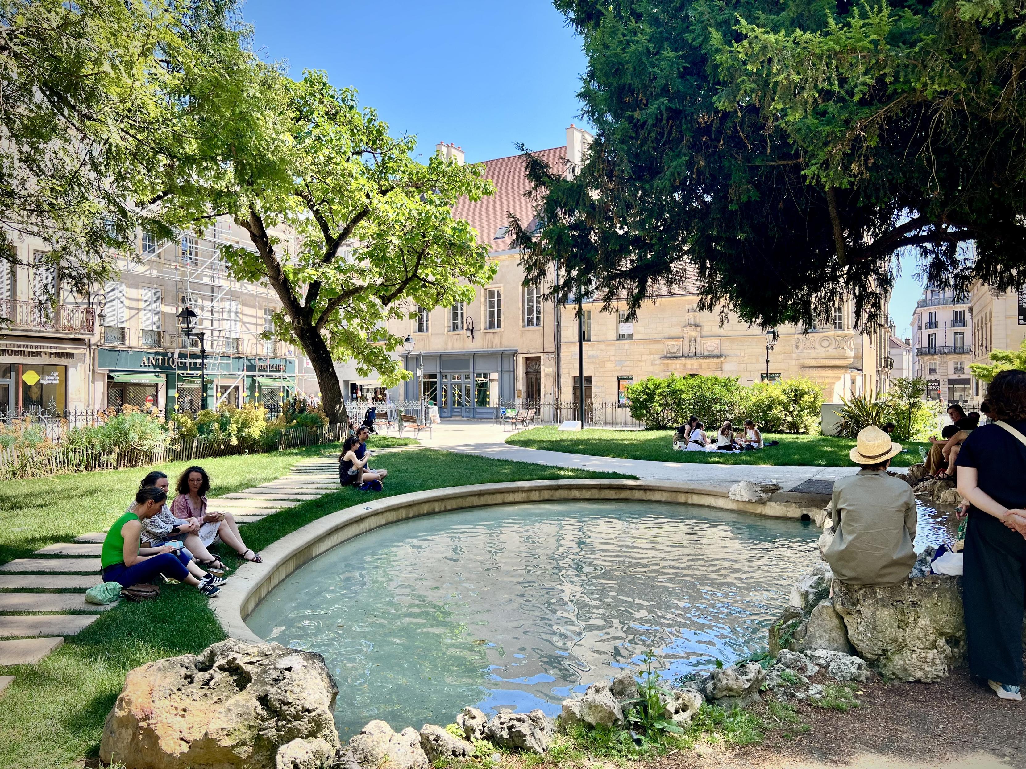 Place des Ducs de Bourgogne, Dijon

A bright outdoor courtyard garden in a sunny day. In the foreground, a small circular stone fountain holds clear, gently rippling water. The surrounding area is covered in vivid green grass and trimmed hedges, with rocks placed along the edge of the fountain. Curving stone paths lead through the garden.

People sit on the grass and along the walkway, some resting in small groups. Tall leafy trees fill the left and right sides, casting dappled shade. Behind the garden, pale buildings with many windows and a red-tiled roof form the background, and a few shops with awnings line the left side.