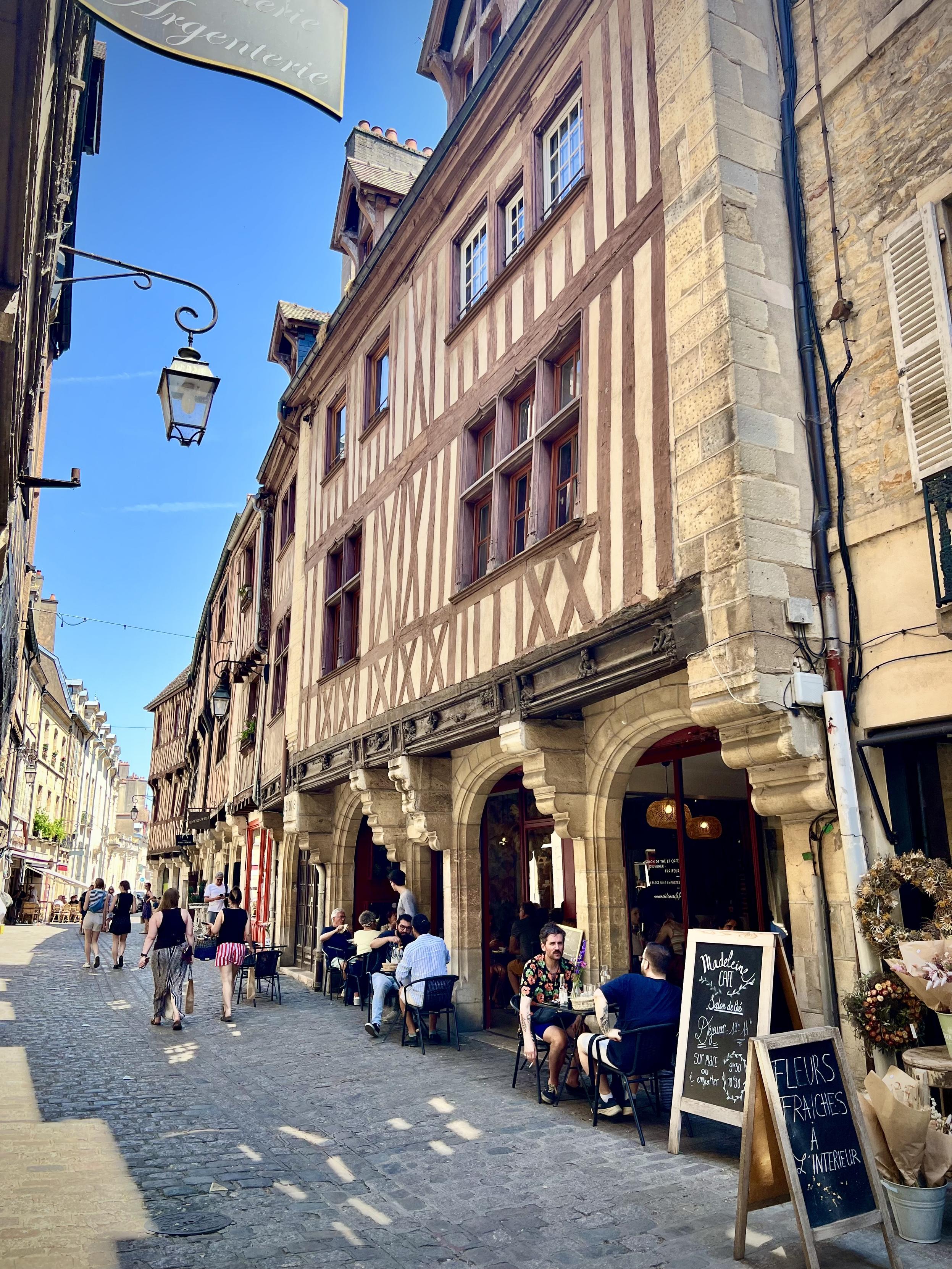 Rue Verrerie, Dijon

A lively street scene in a historic French town. The camera looks down a narrow cobblestone lane lined with half-timbered buildings—cream walls with reddish-brown wooden beams—and tall, arched stone storefront entrances at street level. Sunlight falls from above, creating crisp shadows along the façades.

In the foreground right, an outdoor café seating area occupies the sidewalk with dark chairs and people gathered around small tables. A blackboard menu stand and a flower display add color near the entrance. Further down the street, more pedestrians stroll along the lane under a clear blue sky, with traditional lanterns and hanging shop signs visible along the left side.