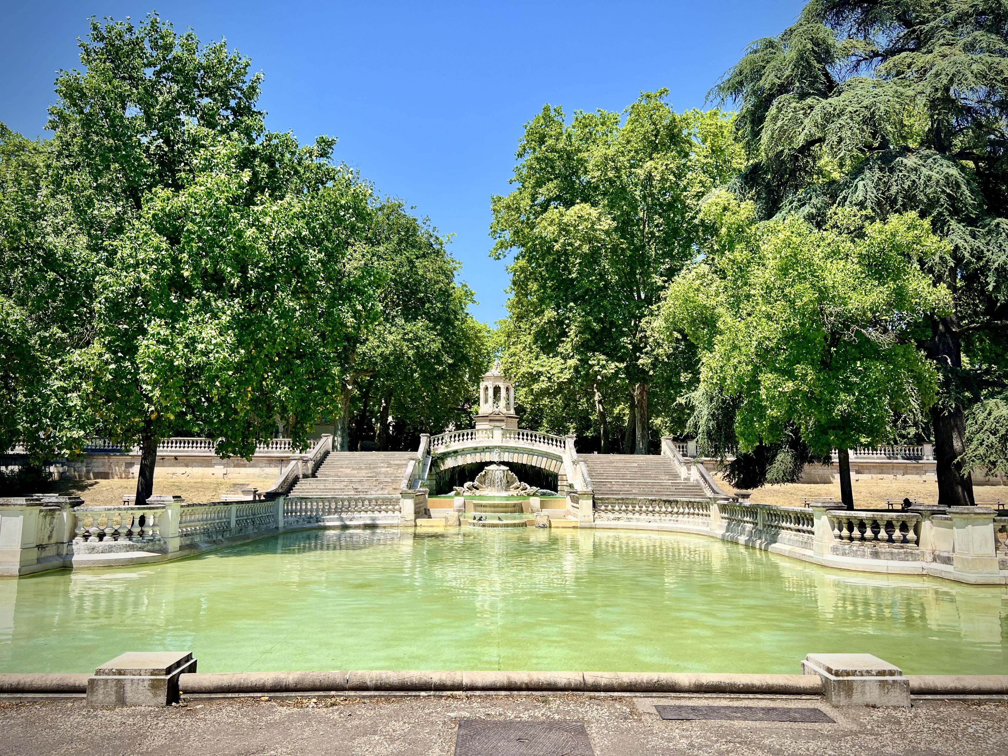 Jardin Darcy, Dijon

A tranquil park fountain scene in bright daylight. The foreground is dominated by a wide, greenish pool of water, surrounded by pale stone balustrades and rounded steps. A larger central feature—an arched stone bridge over the fountain—sits behind the water, with steps rising on both sides.

The background is framed by tall, leafy trees with dense green canopies on both the left and right, creating a natural, sheltered atmosphere. In the distance, more stonework and a small structure rise above the fountain area under a clear blue sky.