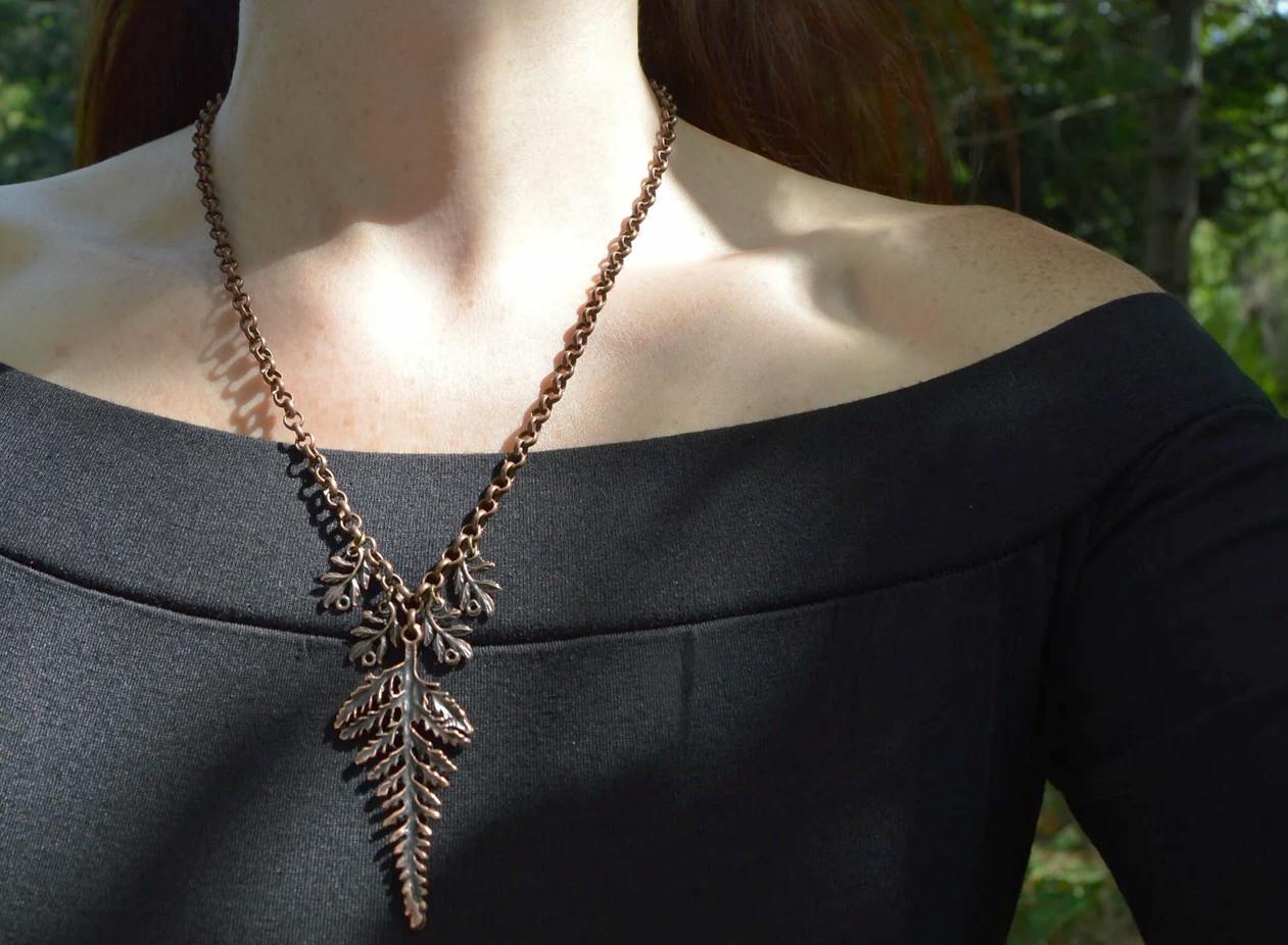 Copper fern leaf pendant on a long chain. Worn by a figure in black standing in a dark forest. Smaller copper fern leaves hand either side of the main fern pendant.