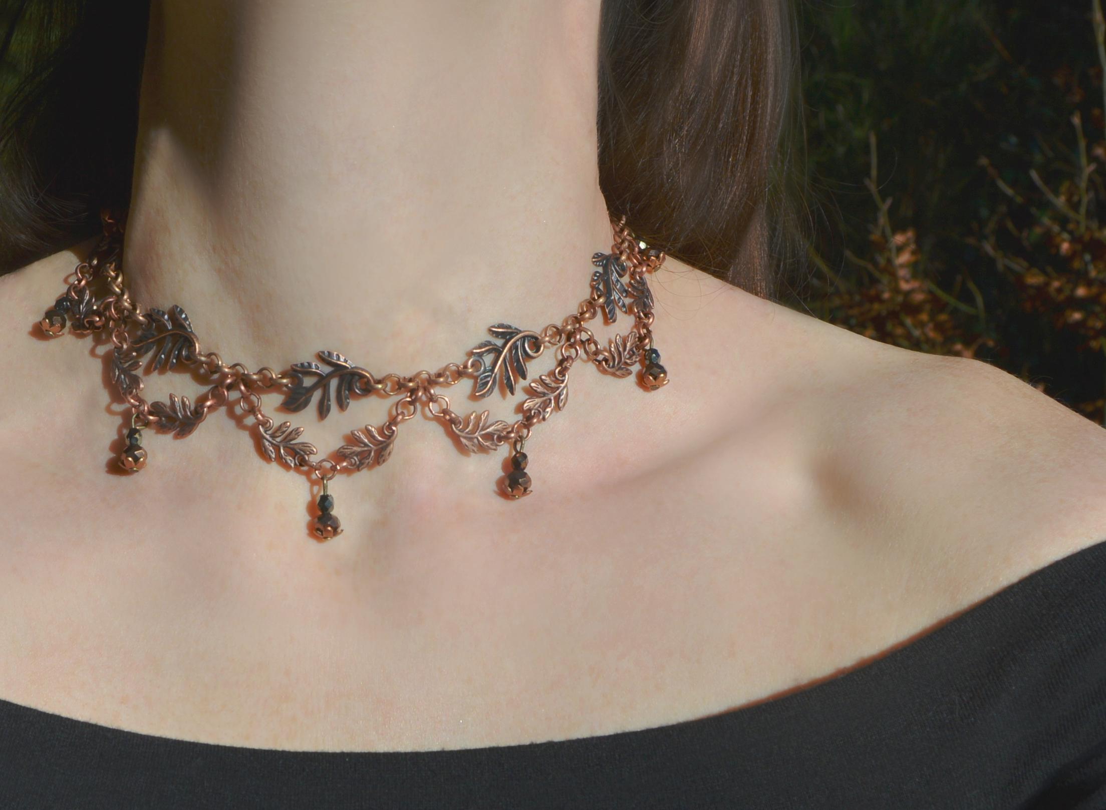Choker necklace of copper fern leaves. Large leaves are linked together by a chain. Smaller fern leaves hang in pairs in v shapes underneath, with tiny bronze beaded pendants hanging from the lowest points. Worn by a pale figure standing in a forest.