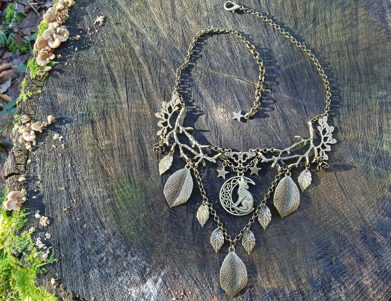 Bronze V shaped necklace made of twigs and leaves, laid flat on a tree stump and viewed from above. Branches form the upper part, from which hang leaves, a howling wolf and moon, and tiny stars.