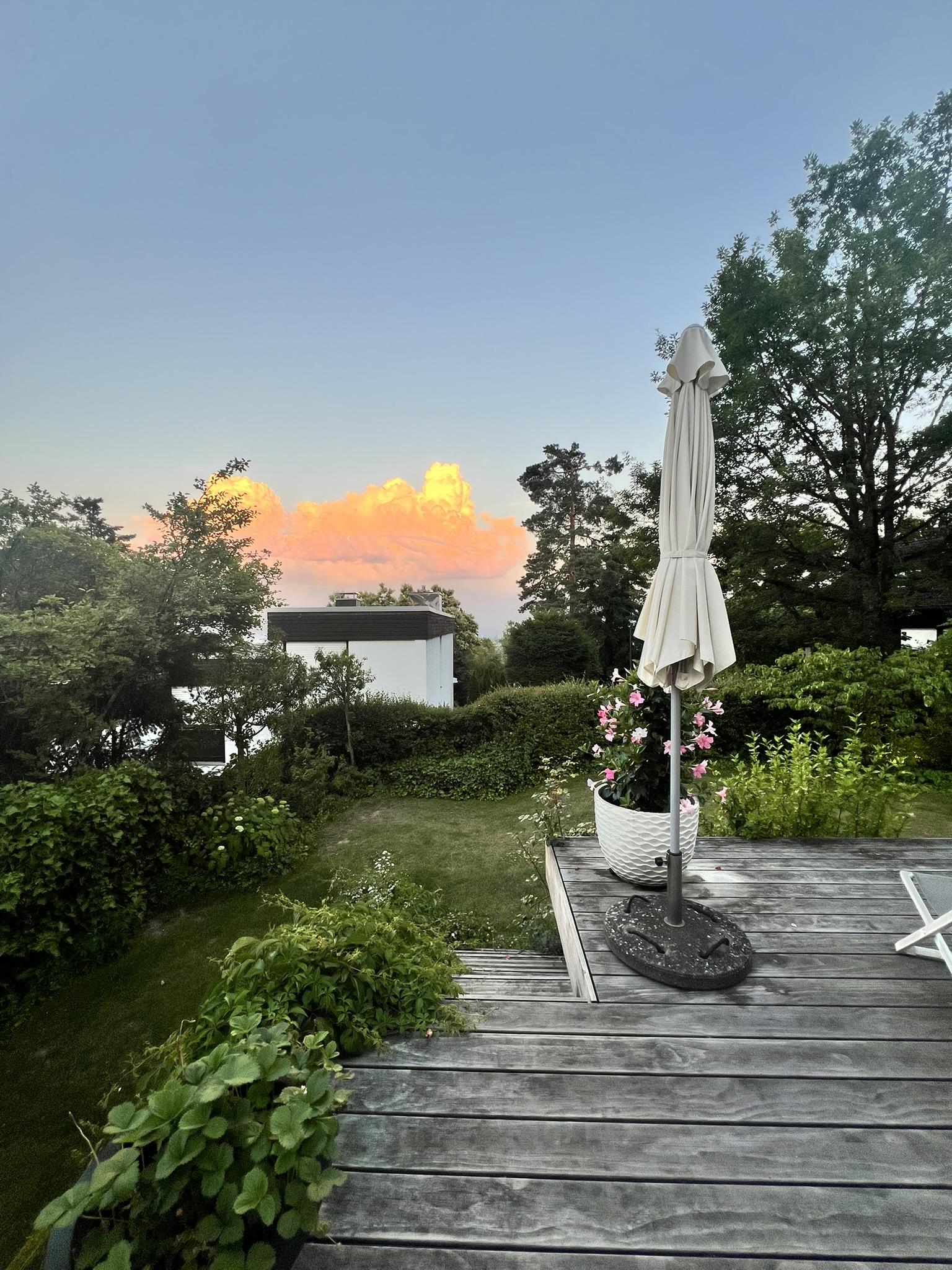 Somewhere in Mammolshain 😉

A wooden deck extends into a green garden, with a folded white patio umbrella in a large pot at the foreground. A modern white house with dark trim sits beyond hedges and trees. A clear blue sky with soft light gives a calm, late-afternoon atmosphere.