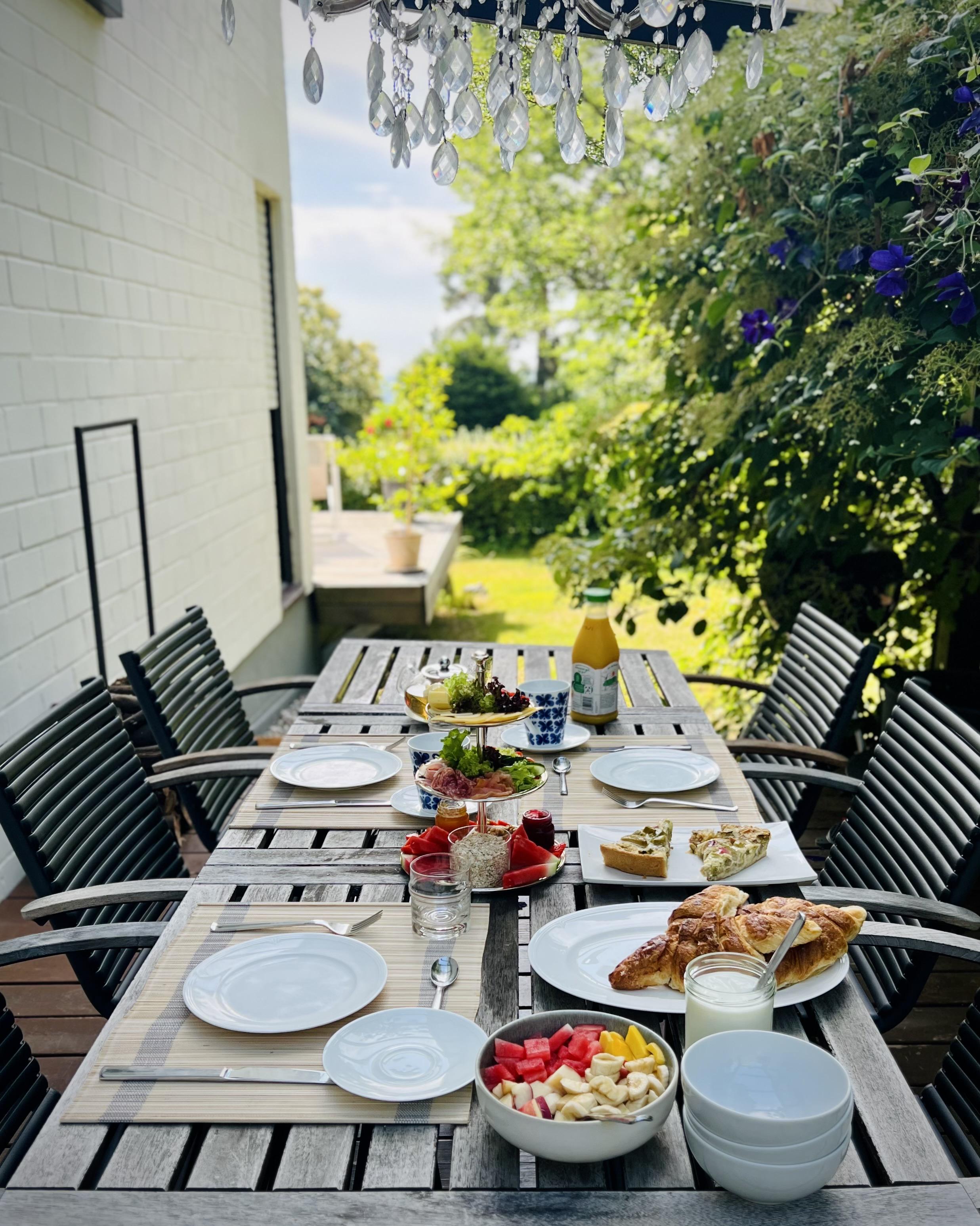 Mammolshain, Frankfurt, Germany

An intimate outdoor dining setup on a narrow patio. The camera looks straight down the long wooden table, surrounded by dark slatted chairs. Plates, cutlery, and glassware are neatly arranged, with bowls of salad and colorful fruit and vegetables, plus bread on one side. Small condiment bottles and a jar sit near the center.

Above, a crystal-like chandelier of hanging clear pendants adds a decorative touch. On the right, dense green foliage climbs along the wall, dotted with blue flowers. A white exterior wall runs along the left, and deeper in the background a bench and a potted plant sit in the sunlit garden space.