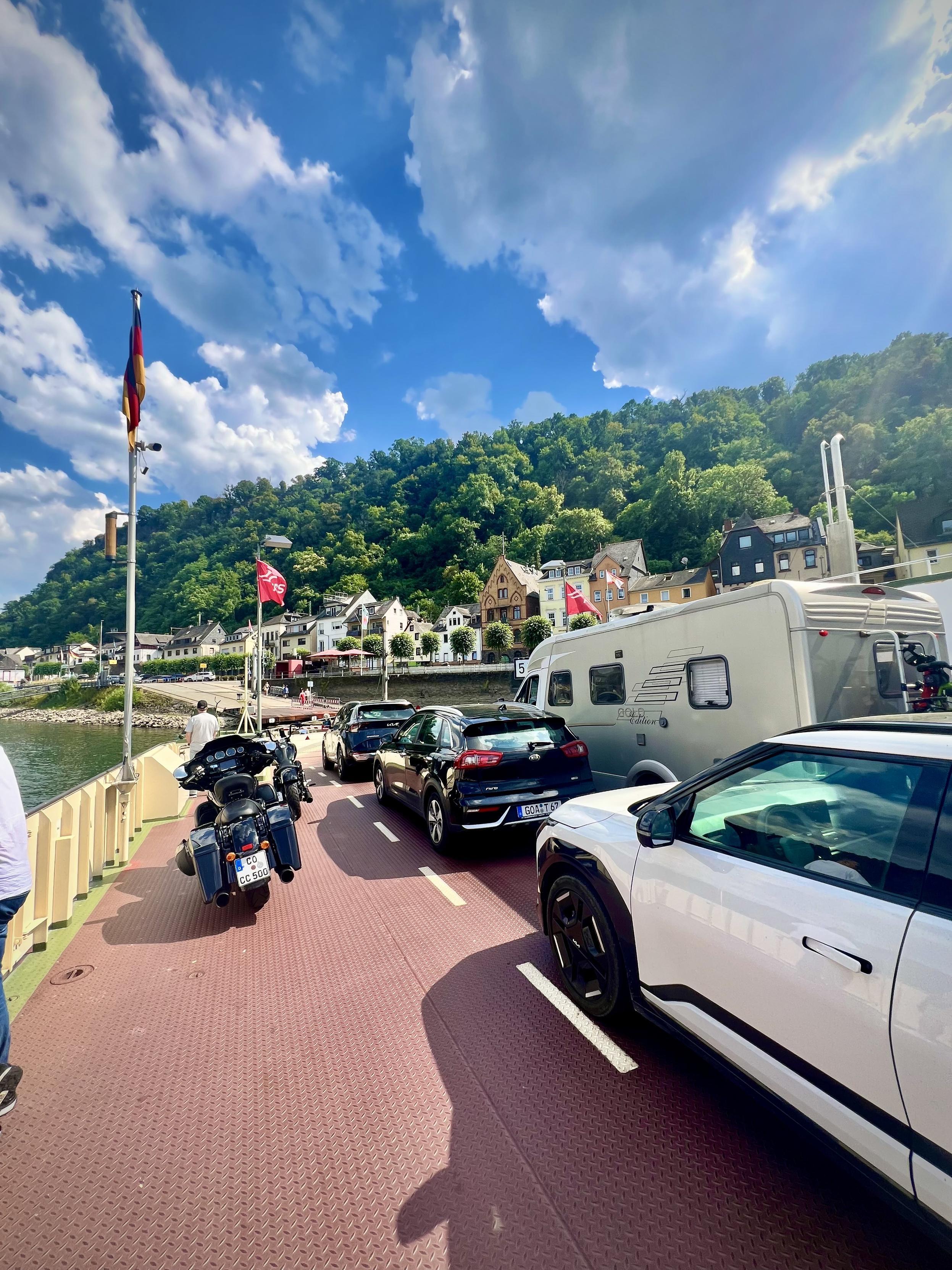 A sunny riverfront parking area with a row of cars and motorcycles. In the foreground right, a white car occupies the frame, while across the lane a large white camper van is parked. On the left, a motorbike stands near a low concrete wall that runs alongside the water.

In the middle distance, a small town lies along the riverbank with colorful houses—white, beige, and pastel—plus a few storefronts and outdoor seating under red umbrellas. Several red flags on tall poles line the walkway.

Behind the town, a steep hillside is covered in dense green trees, rising up toward the top of the frame. Above it all, the sky is bright blue with large, fluffy white clouds.