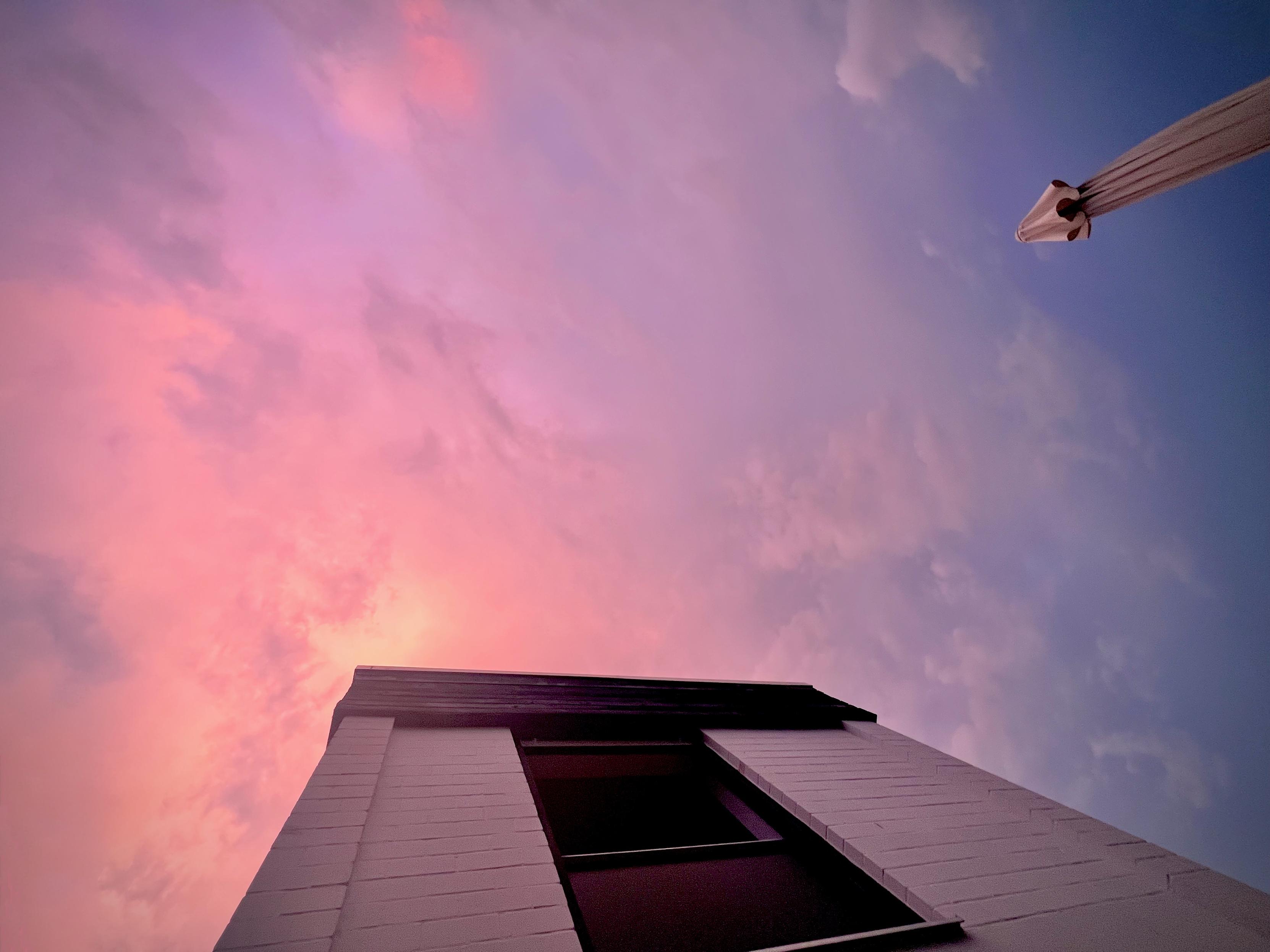 View from the ground up at a tall white-brick building with a dark opening along the side. A slender street lamp extends diagonally from the top-right into the sky. The sky shows a gradient from pinkish hues on the left to blue on the right, with soft clouds.