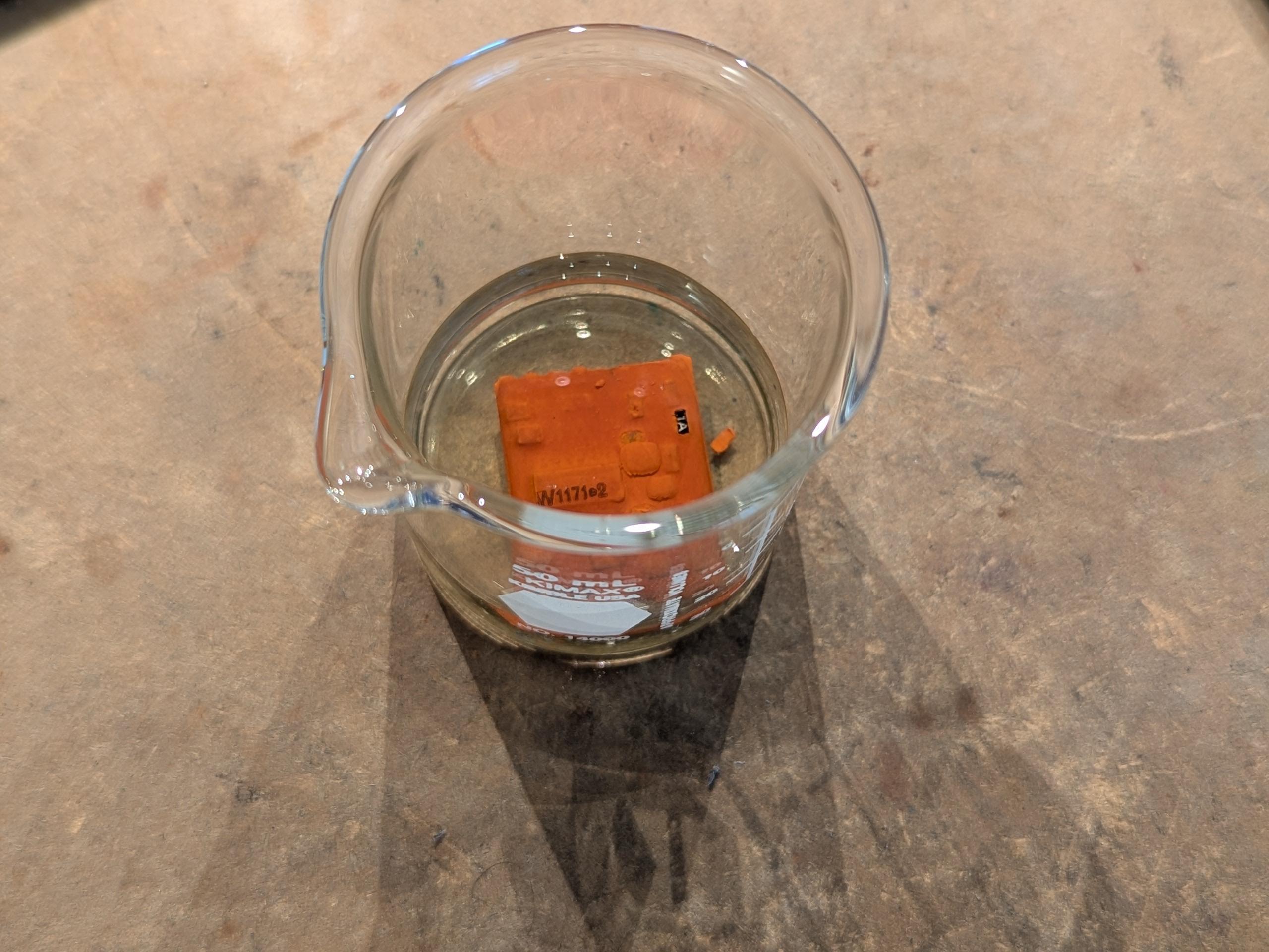 the module submerged in acetone inside a tiny glass breaker. part of the brown potting compound has fallen off revealing a sot-23 transistor marked "1A".