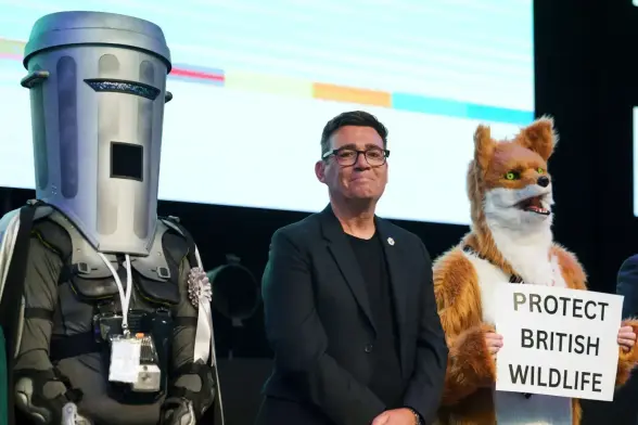 Person in trashcan costume and another an a rather ugly fox fursuit holding a sign that says PROTECT BRITISH WILDLIFE flank a politician who is almost smiling in front of a projection screen