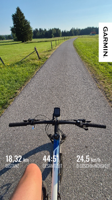 This image captures a first-person perspective of a cycling ride on a scenic, paved path through a lush, green landscape. The view is from the cyclist's handlebars, with the road stretching ahead into the distance, flanked by vibrant green meadows and a line of evergreen trees. The path is smooth and well-maintained, inviting a sense of freedom and adventure.

The cyclist's legs are visible at the bottom of the image, and the handlebars and a mounted device (likely a GPS or cycling computer) ar…