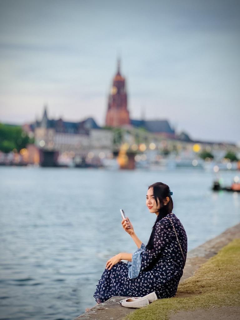 A woman in a dark blue floral dress sits on a low stone ledge beside a calm river, with her legs folded and hands resting on her knee. She holds a smartphone in her right hand and looks to the side with a gentle smile; a white sneaker is near her foot. In the blurred background, a city skyline across the water features a tall brick tower. The scene is softly lit, conveying a tranquil, contemplative mood.