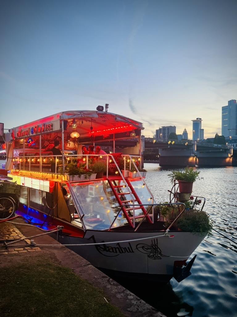 A double-decker boat named "Istanbul" is docked along a riverbank at twilight, brightly illuminated with red neon and warm string lights. The boat serves as an outdoor dining spot, featuring potted plants on deck and patrons seated under a canopy. In the background, a bridge crosses the water, leading toward a modern city skyline under a fading evening sky.